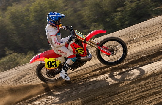 A person riding a red dirt bike uphill, leaving a trail of dust behind