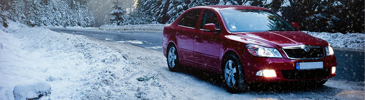 A red compact car parked on the side of a snowy road, with snow-covered pine trees in the background