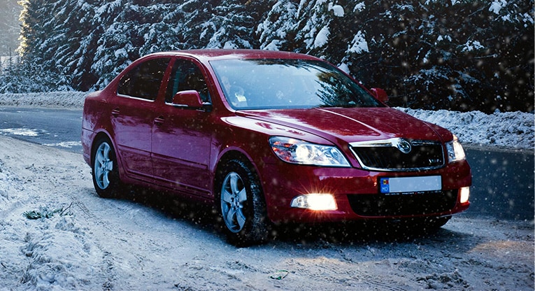 A red compact car parked on the side of a snowy road, with snow-covered pine trees in the background