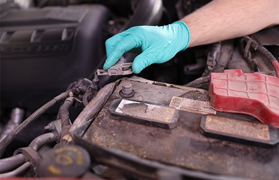 Close-up of a person wearing a green nitrile glove disconnecting a terminal from a dusty, corroded car battery under a vehicle hood.
